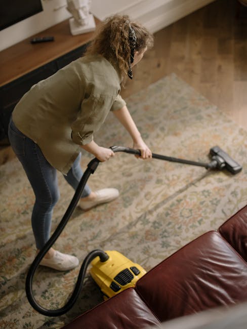 A person using a yellow and black vacuum cleaner on a patterned area rug, with their hands placing a cleaning cloth into the machine's compartment. The room has a wooden floor visible in the backgroun