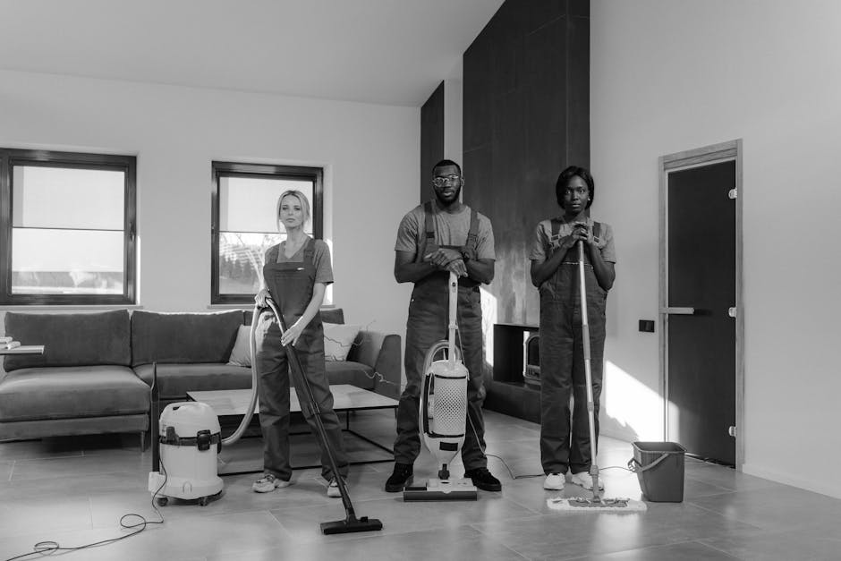 A team of three professional cleaners, dressed in dark uniforms with aprons, are performing surface cleaning and deep cleaning in a modern living room. The room features a large sectional sofa upholst